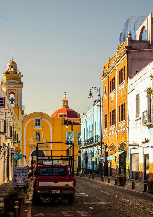 Una camioneta roja aparcada en una calle estrecha flanqueada por edificios coloridos, que conduce hacia una iglesia amarilla con cúpula roja.