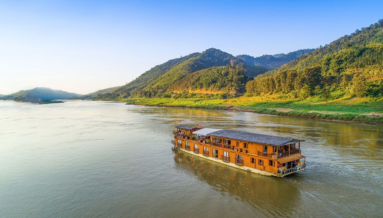 Una barca di legno a due piani naviga su un ampio fiume, affiancata da rigogliose colline verdi sotto un cielo azzurro chiaro.