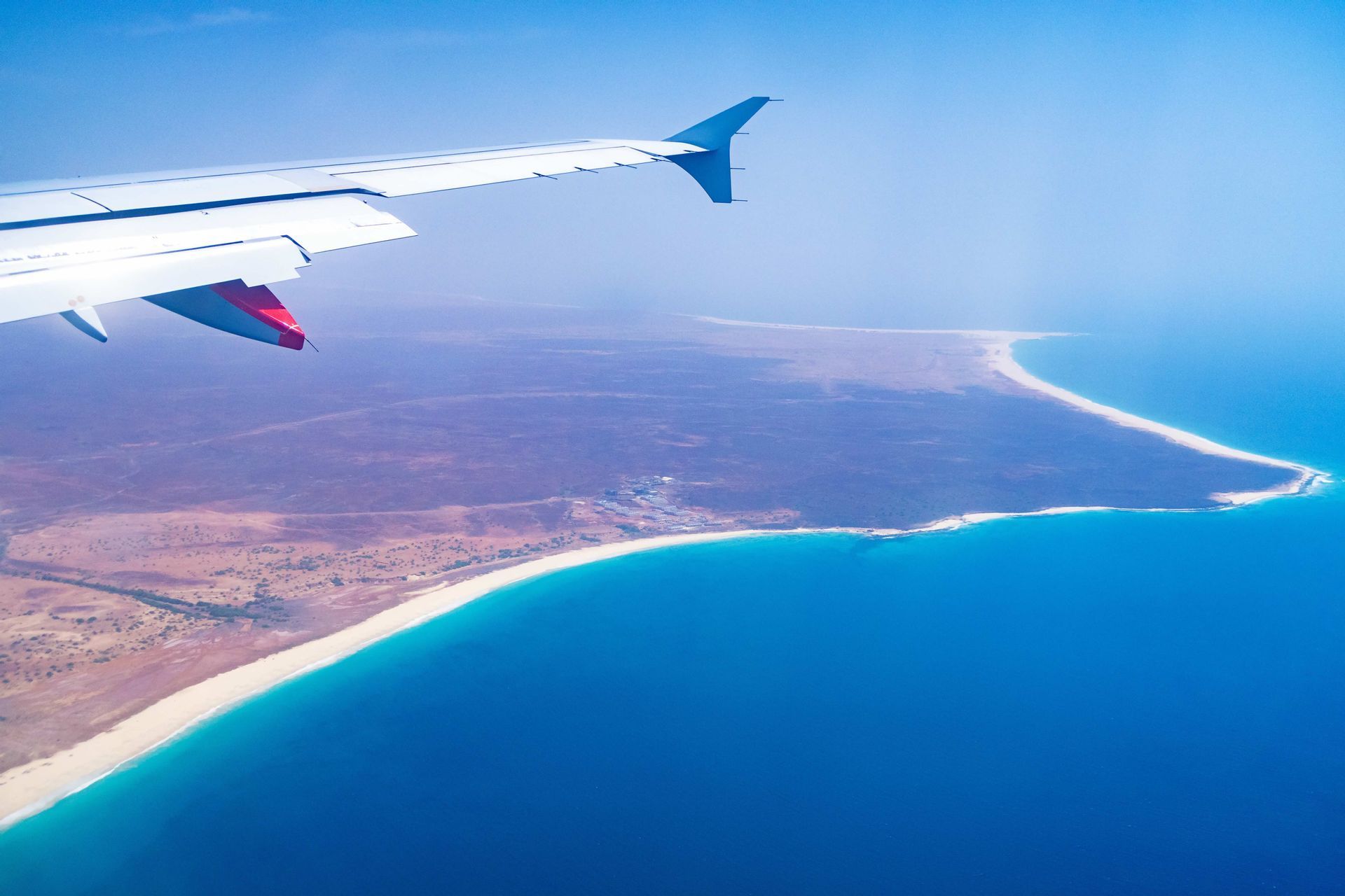 Une aile d'avion vue depuis un hublot, survolant un littoral tropical avec une plage de sable blanc et une mer turquoise.