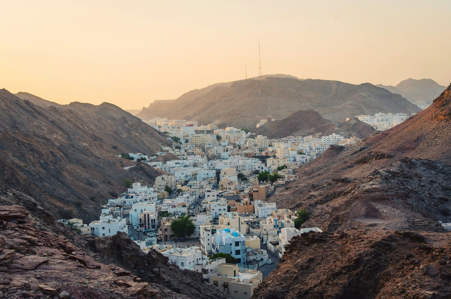 A town of white buildings nestled in a rocky valley surrounded by mountains under a hazy golden sky.