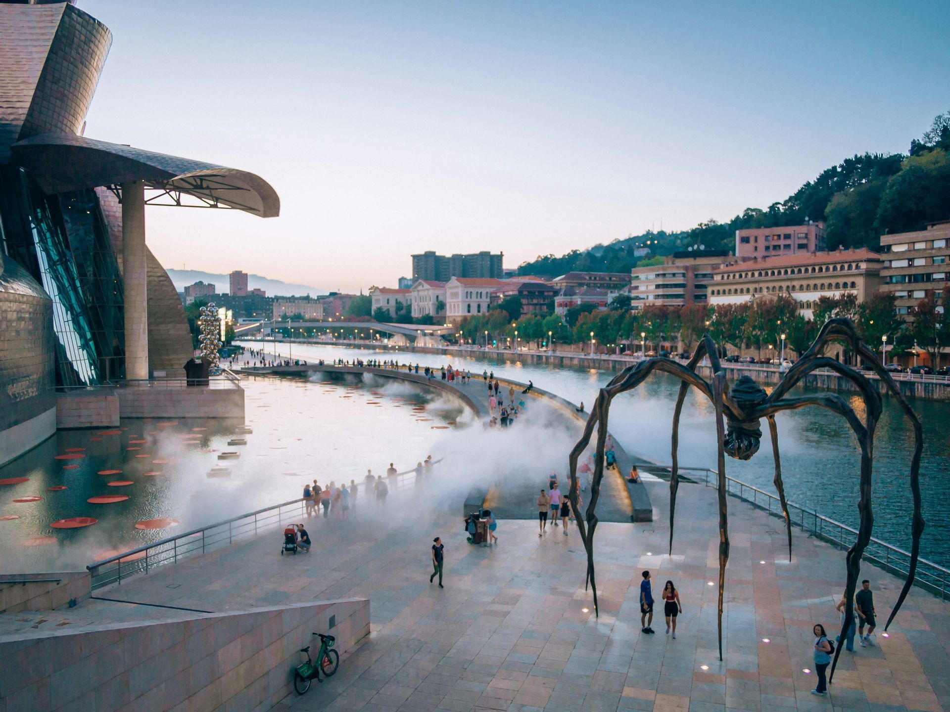 Una grande scultura di ragno in metallo su una passeggiata lungofiume accanto a un edificio moderno al crepuscolo, con persone che camminano nella nebbia.