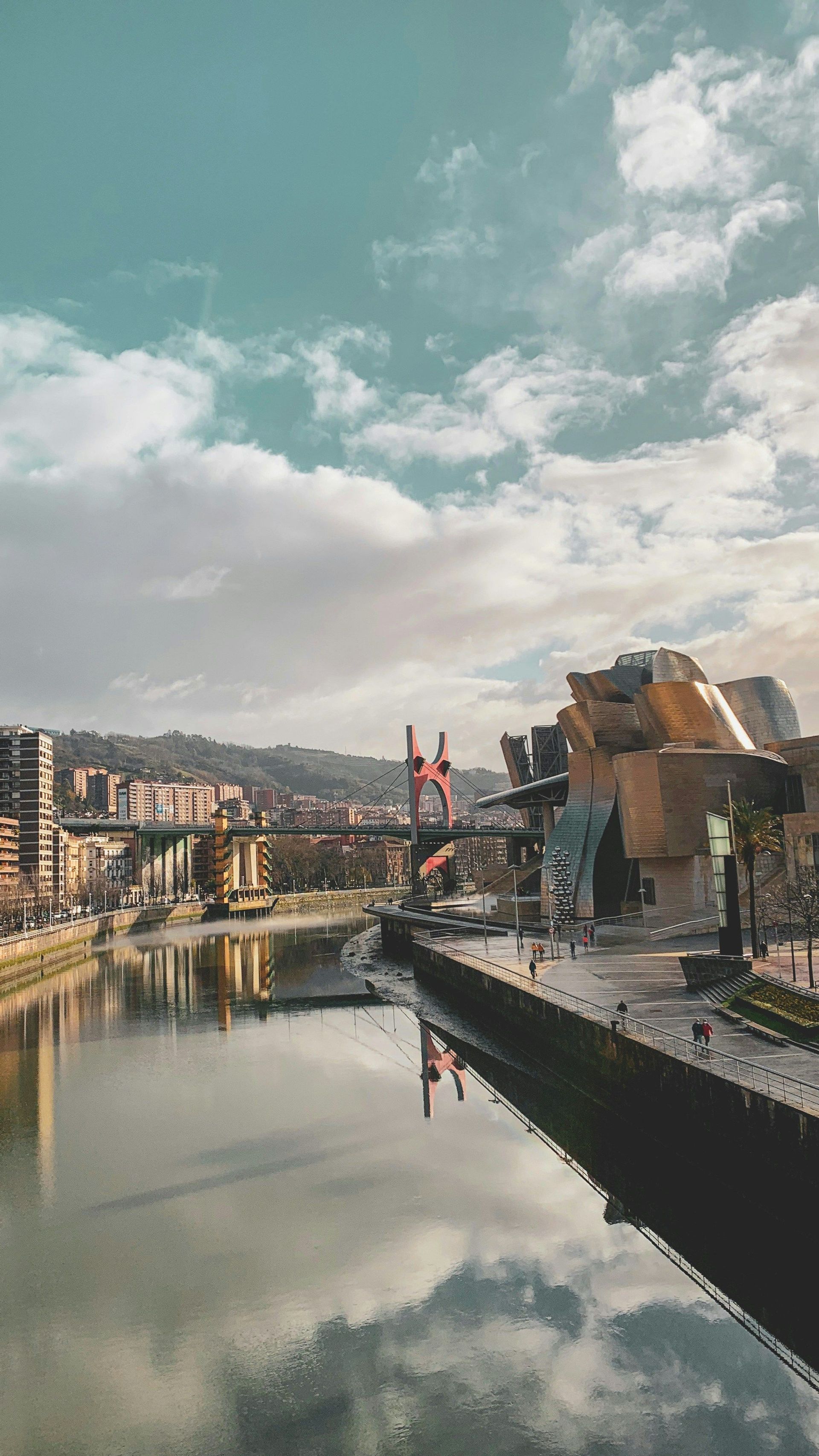 Un moderno edificio architettonico su una riva di fiume con il suo riflesso nell'acqua, un ponte rosso e una città sullo sfondo.