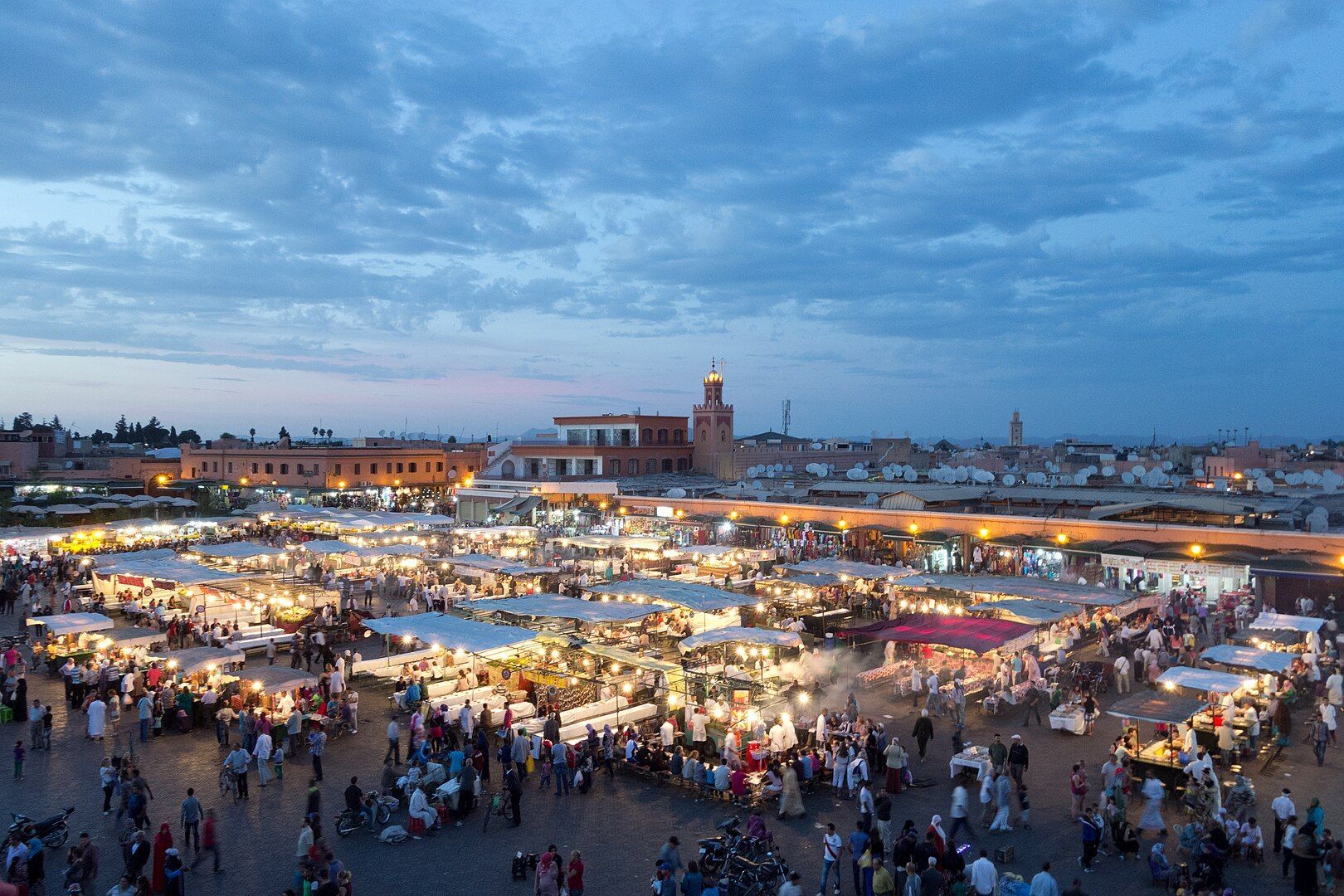 Una vista dall'alto di una vivace piazza del mercato all'aperto al tramonto, affollata di gente e bancarelle di cibo illuminate a festa sotto un cielo nuvoloso.