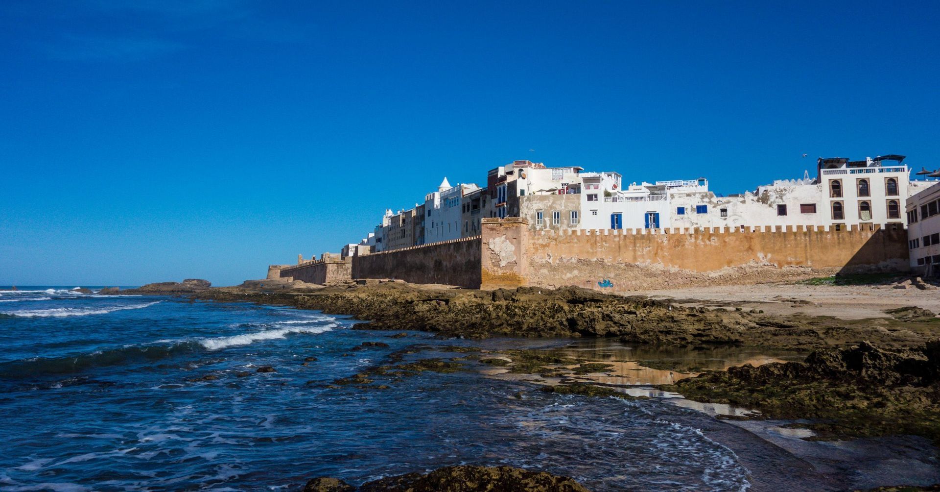 Una città fortificata con edifici bianchi si erge su un muro di cinta logoro su una costa rocciosa, con le onde azzurre dell'oceano che si infrangono sulla riva.