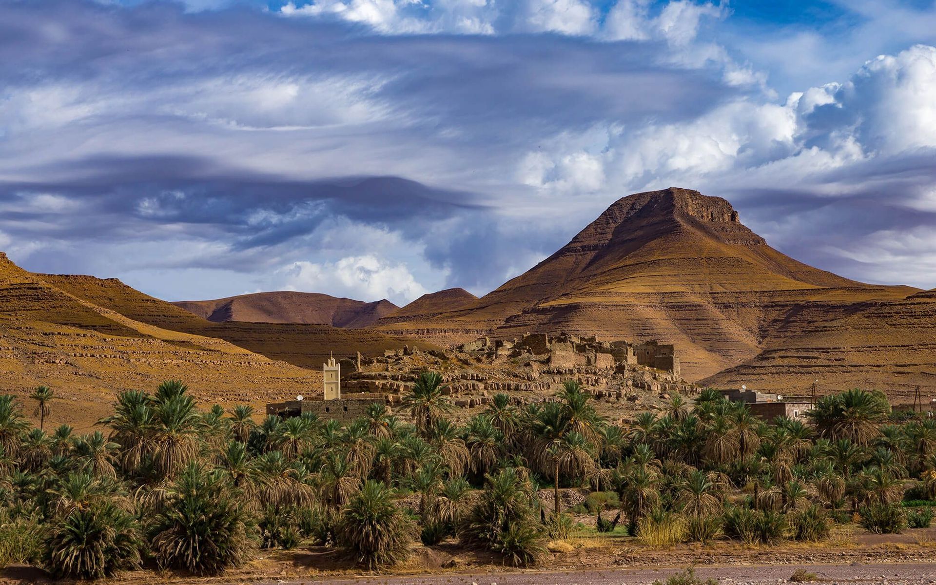Un antico villaggio e minareto incastonati in una lussureggiante oasi di palme, ai piedi di aride montagne stratificate sotto un cielo nuvoloso.
