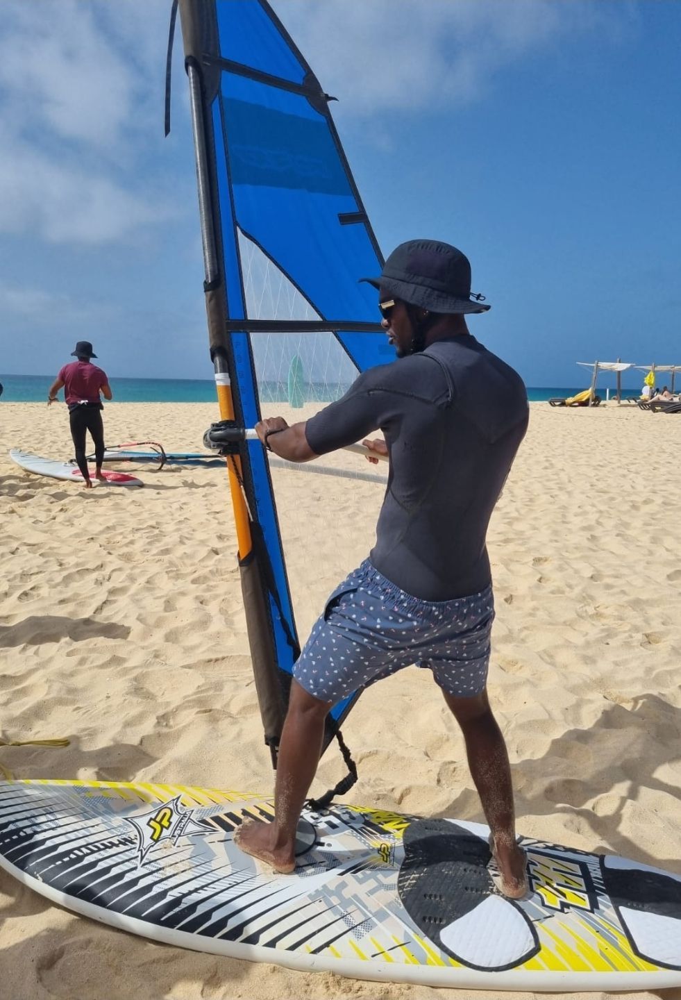 Un uomo con la parte superiore di una muta e un cappello da pescatore sta su una tavola da windsurf su una spiaggia sabbiosa, tenendo il boma della vela.