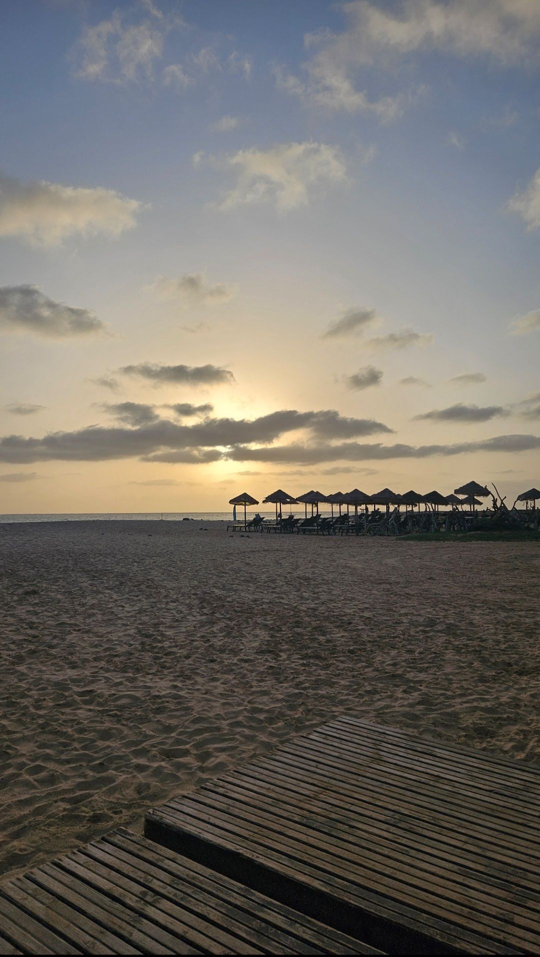 Una passerella di legno su una spiaggia sabbiosa, con una fila di ombrelloni e lettini sagomati contro il tramonto sull'oceano.