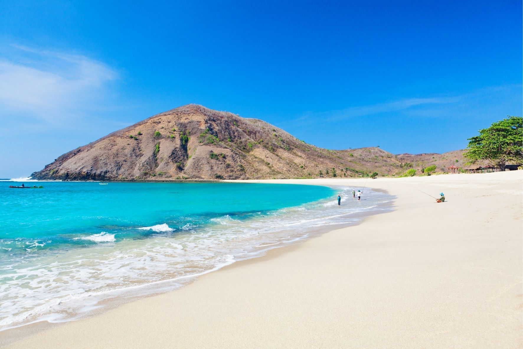 Ein weißer Sandstrand grenzt an eine Bucht mit türkisfarbenem Wasser, im Hintergrund ein großer, karger Hügel unter strahlend blauem Himmel.