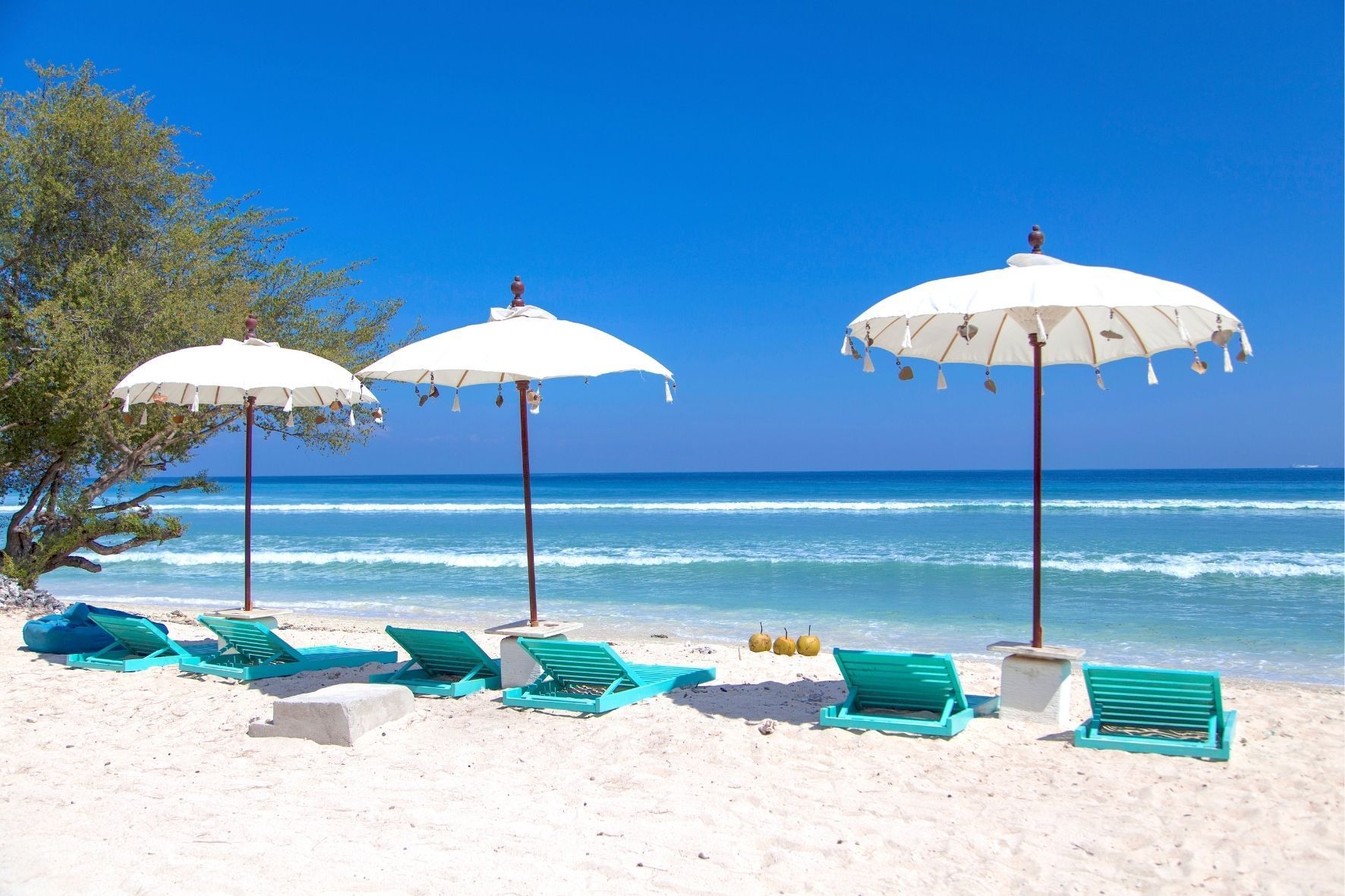 Türkisfarbene Liegestühle und weiße Sonnenschirme stehen an einem weißen Sandstrand mit Blick auf das türkisfarbene Meer unter einem klaren blauen Himmel.