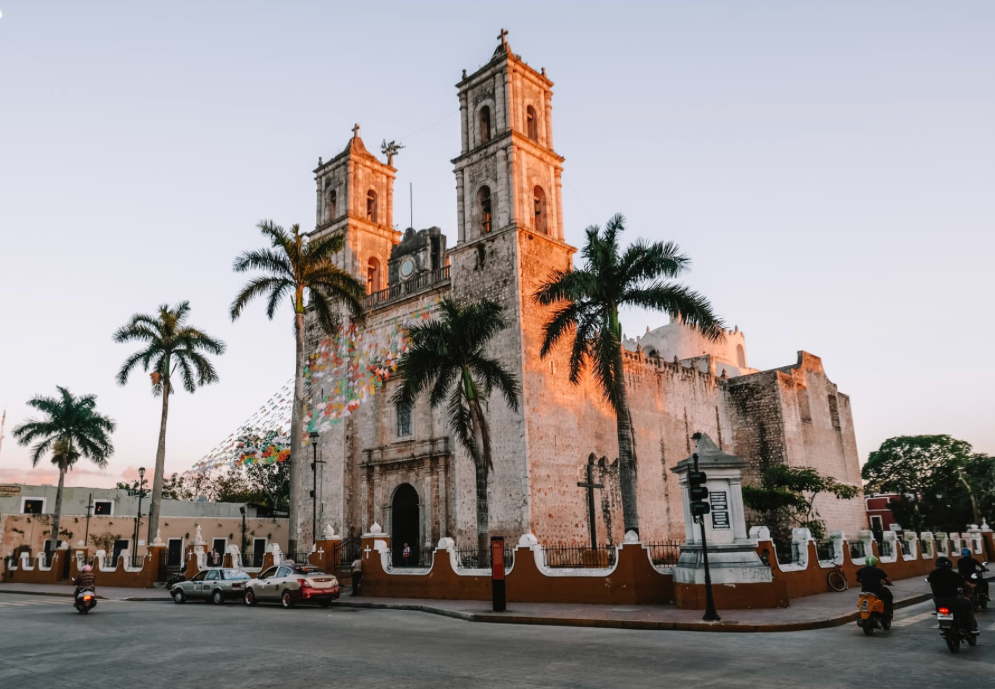 An old stone church with two bell towers is lit by the warm glow of sunset, with palm trees and a street with traffic in the foreground.