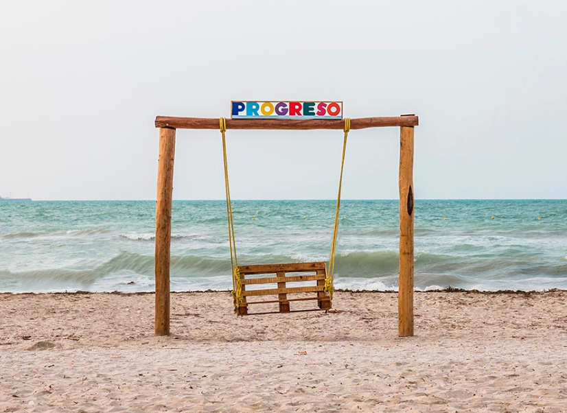 A rustic wooden swing set with a colorful sign on top sits empty on a sandy beach facing the ocean.