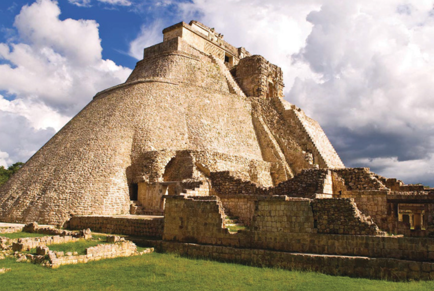 A large, ancient stone pyramid with rounded sides and a steep staircase stands over smaller ruins on a grassy field under a blue sky with white clouds.