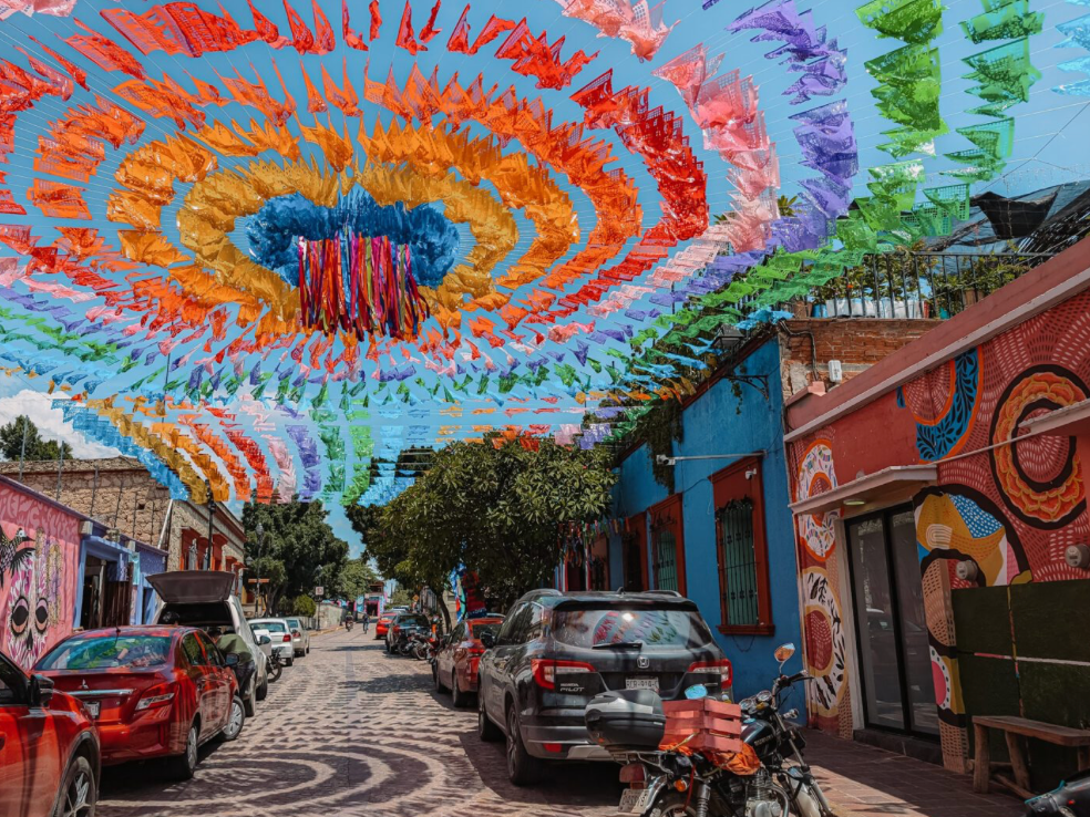 Colorful flags hang in circular patterns over a cobblestone street lined with parked cars and brightly painted buildings.