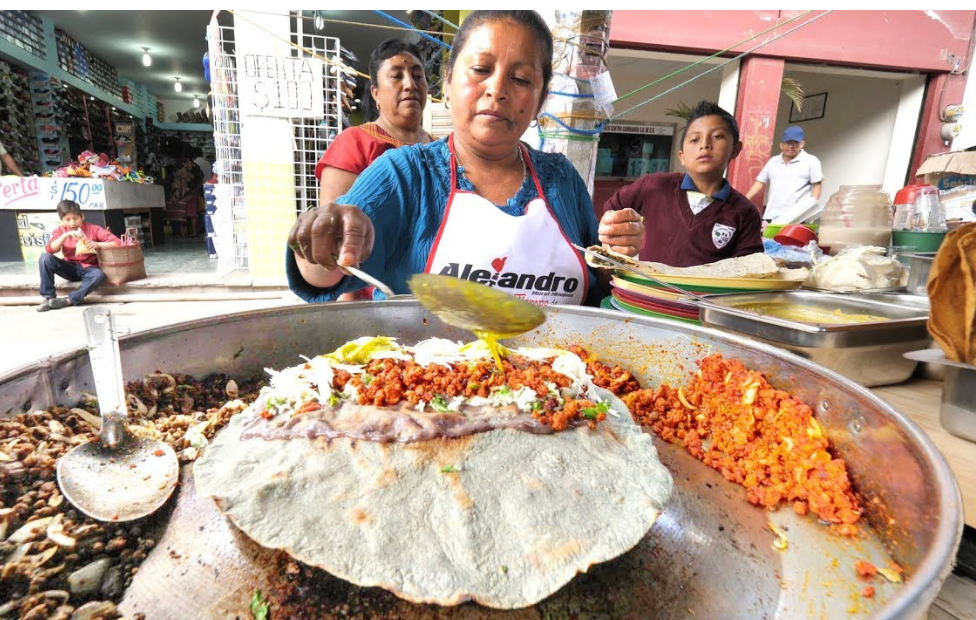 A street food vendor spoons a yellow sauce onto a large blue corn tortilla topped with meat on a circular griddle at a market stall.