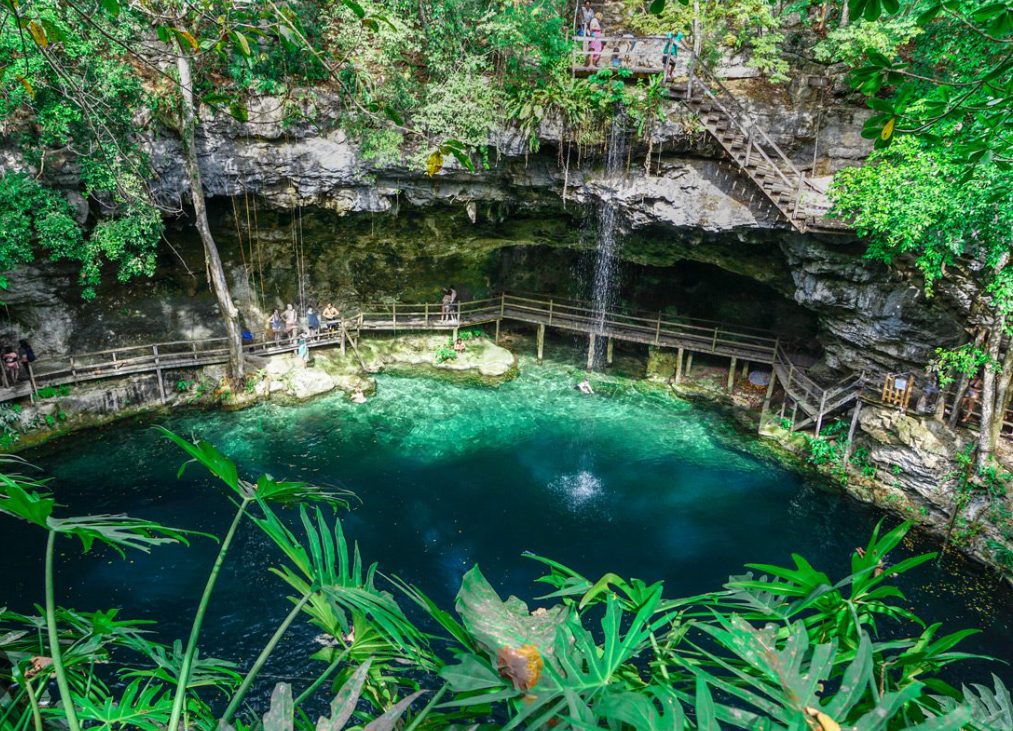 A high-angle view of a WeRoad group trip at a cenote with turquoise water, a waterfall, and wooden walkways built into the rock walls.