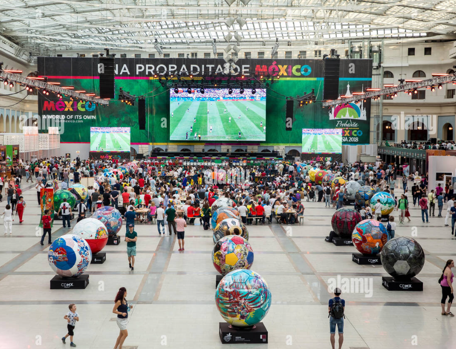 A large crowd gathers in an exhibition hall to watch a soccer match on big screens, with oversized, decorated soccer balls in the foreground.