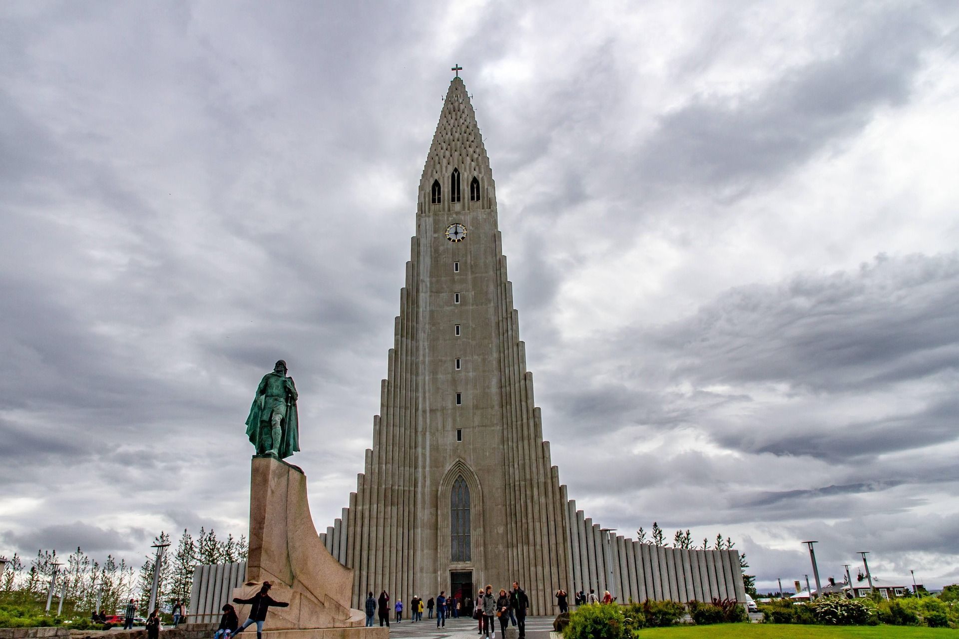 A towering modern church with a stepped concrete facade and a statue on a pedestal in front, under a gray, cloudy sky.