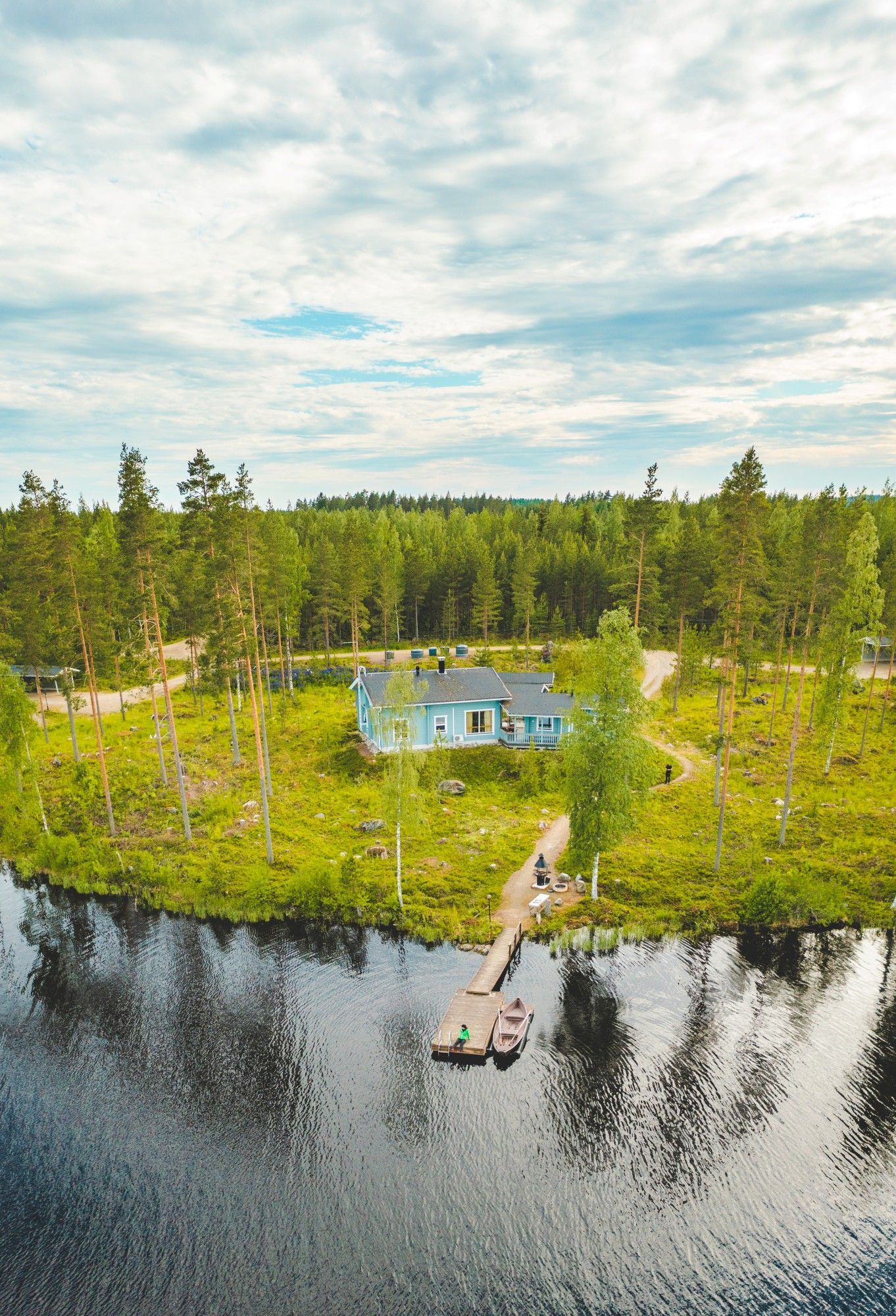 Eine Luftaufnahme eines hellblauen Häuschens auf einer grasbewachsenen Halbinsel, umgeben von einem See und einem dichten Kiefernwald.