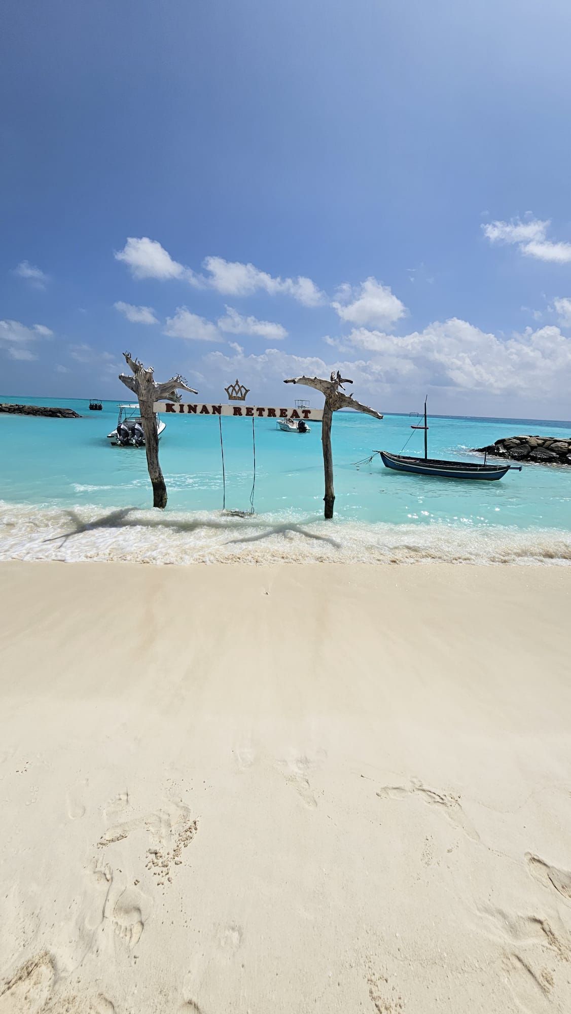 Eine Holzschaukel mit einem 'Kinan Retreat'-Schild steht im flachen türkisfarbenen Wasser eines weißen Sandstrandes unter blauem Himmel.