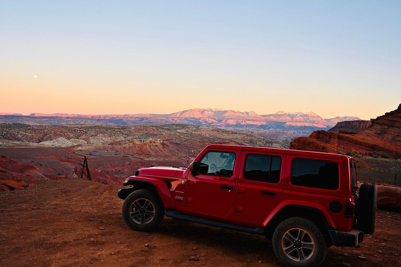 Ein roter Jeep stand auf einem unbefestigten Aussichtspunkt mit Blick auf einen weiten roten Felsencanyon und entfernte, schneebedeckte Berge bei Sonnenuntergang.