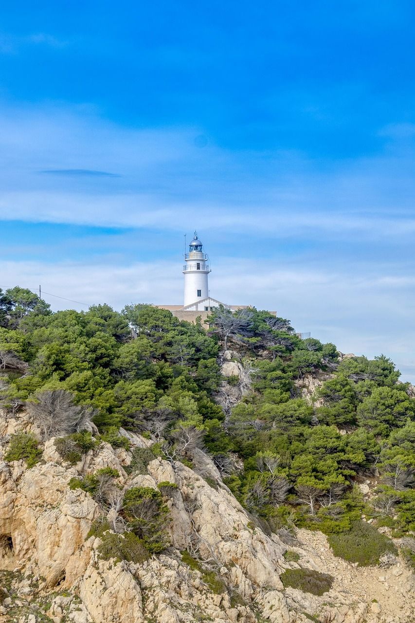 Ein weißer Leuchtturm steht auf einer bewaldeten Felsklippe vor einem strahlend blauen Himmel.