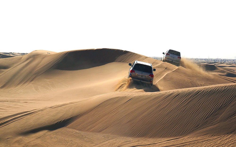 Zwei silberne SUVs fahren über große, rollende Sanddünen in einer Wüste und wirbeln dabei Sand von ihren Reifen auf.