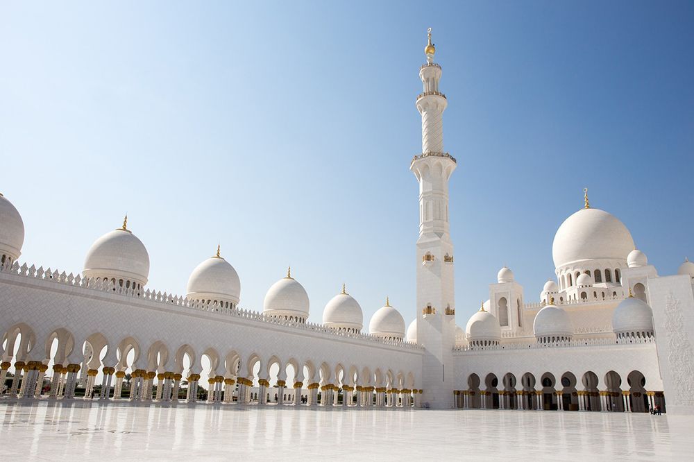 Eine große weiße Moschee mit mehreren Kuppeln, einem Minarett und einem Arkadenhof steht unter einem klaren blauen Himmel.