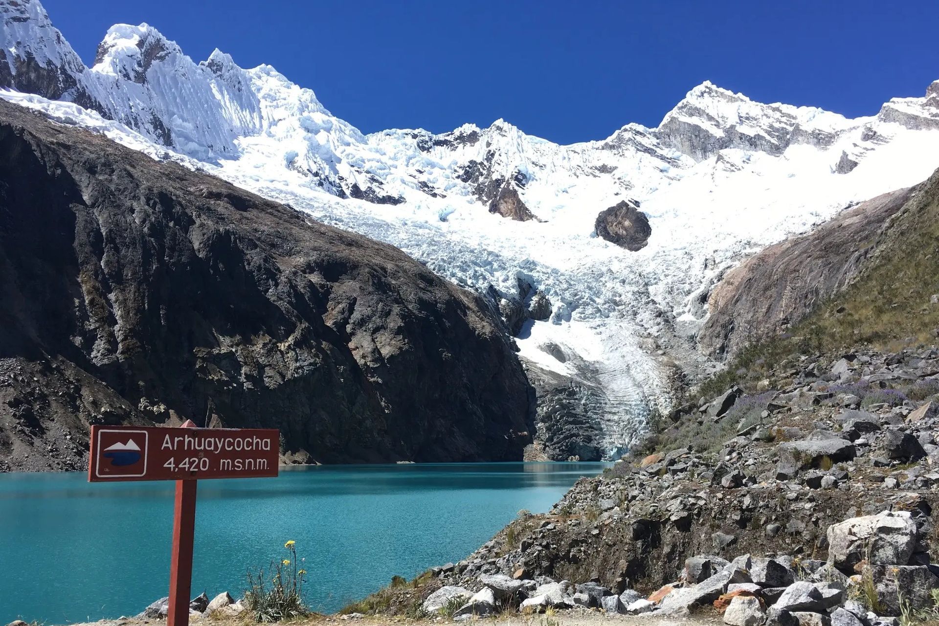 Un cartello di legno per Arhuaycocha accanto a un lago glaciale turchese ai piedi di imponenti montagne innevate e un ghiacciaio.