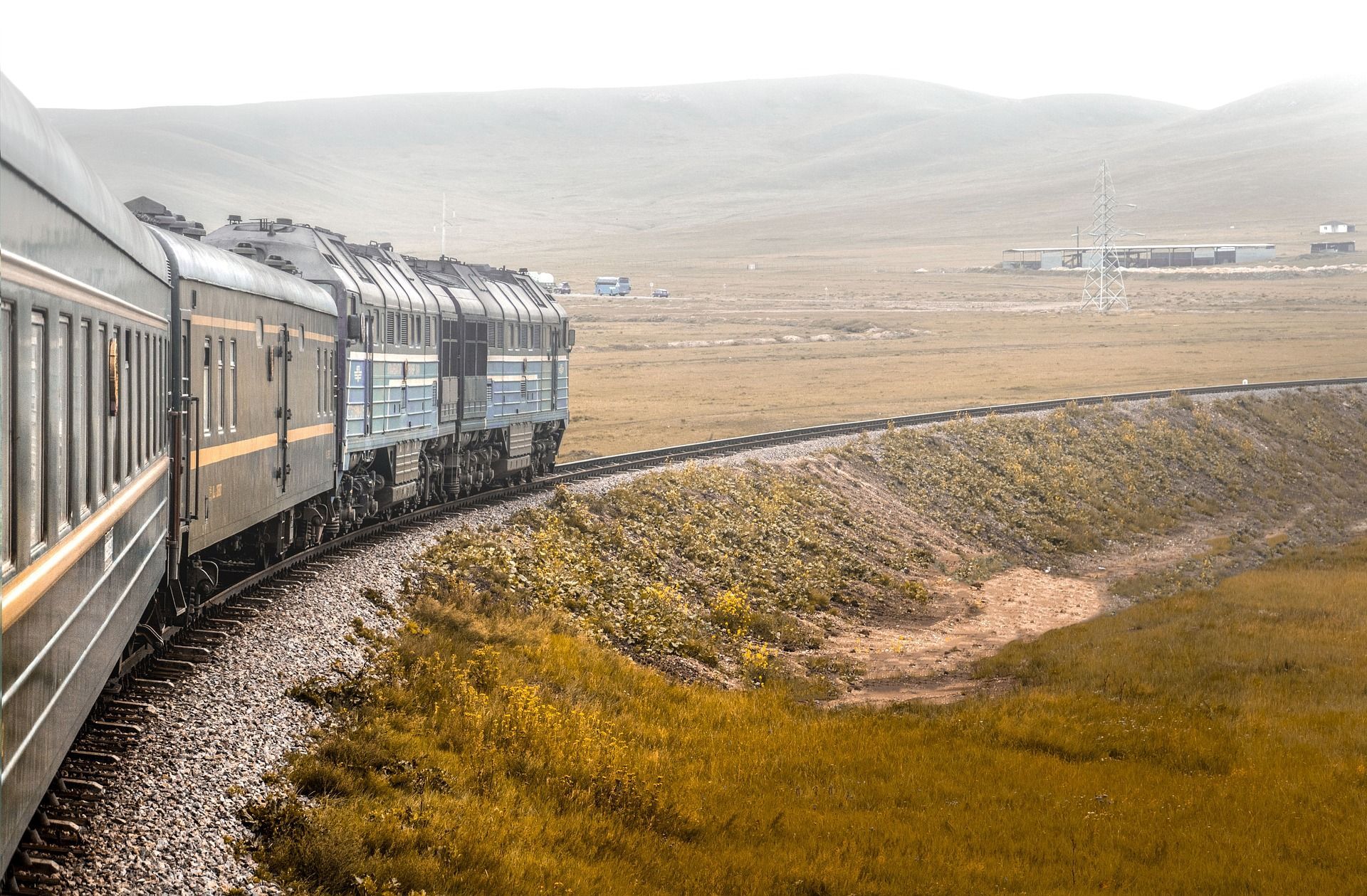 Un lungo treno passeggeri con una locomotiva blu viaggia su un binario curvo attraverso una vasta pianura erbosa con colline nella distanza nebbiosa.