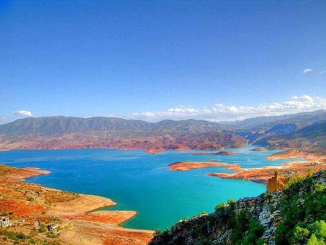Un lago turchese vibrante si snoda tra colline bruno-rossastre con una catena montuosa sullo sfondo sotto un cielo azzurro limpido.
