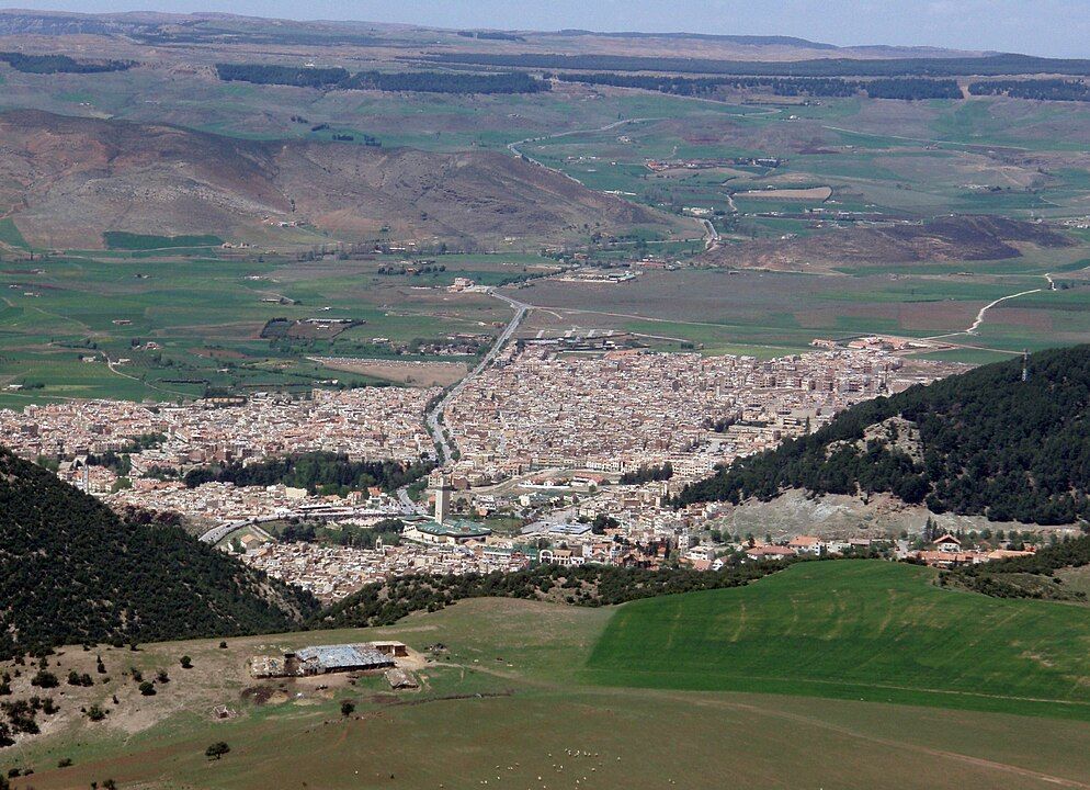 Una veduta aerea di una città tentacolare situata in una valle, circondata da dolci colline verdi e marroni e foreste.