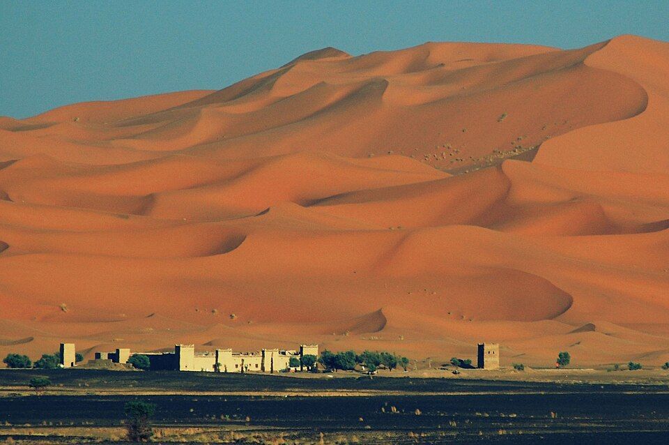 Ai piedi di vaste e ondulate dune di sabbia arancione, sotto un cielo azzurro e limpido, sorge un piccolo villaggio desertico con alberi.