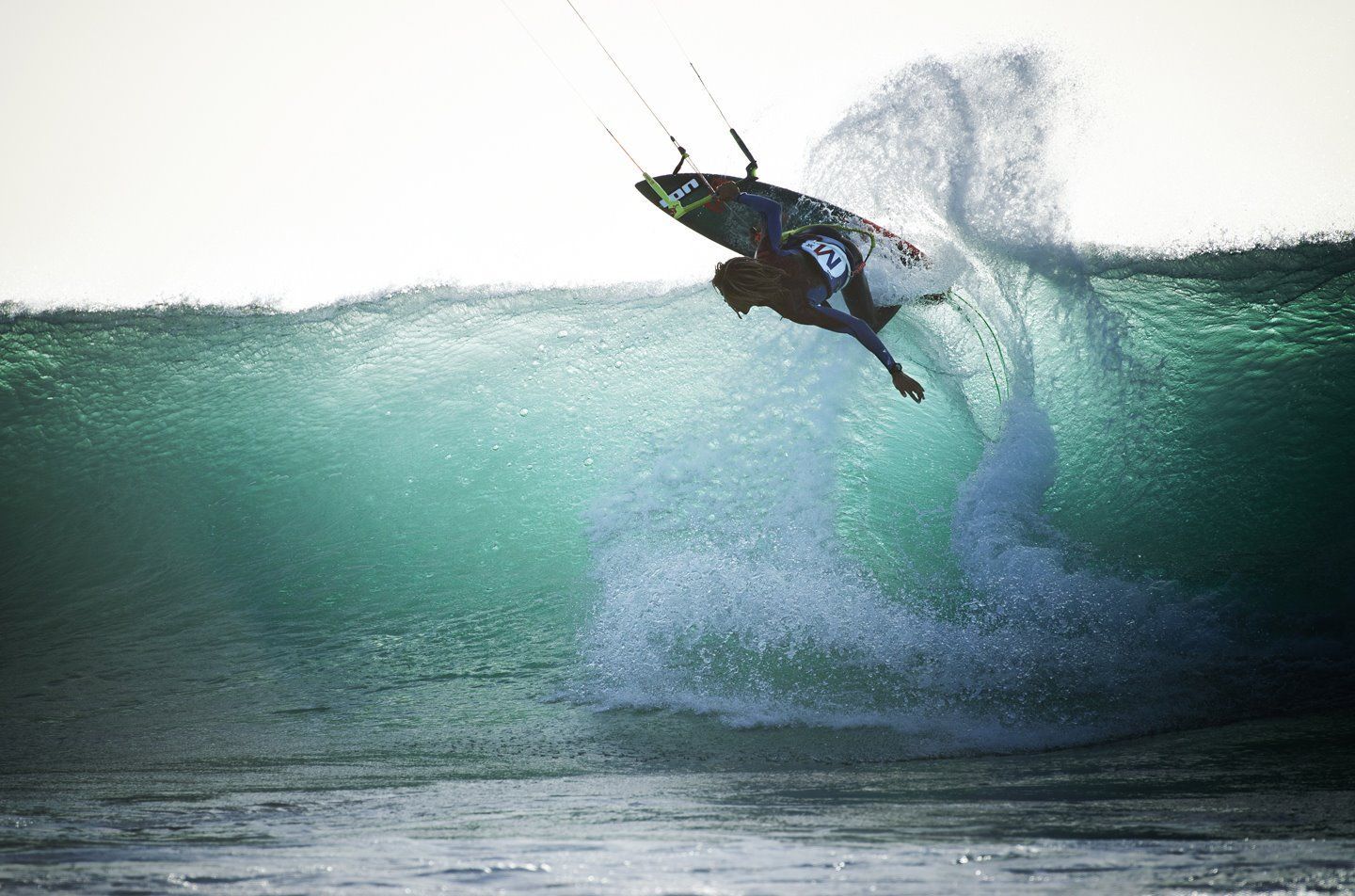 Un kitesurfer dai capelli lunghi manovra lungo la cresta di una grande onda oceanica turchese.