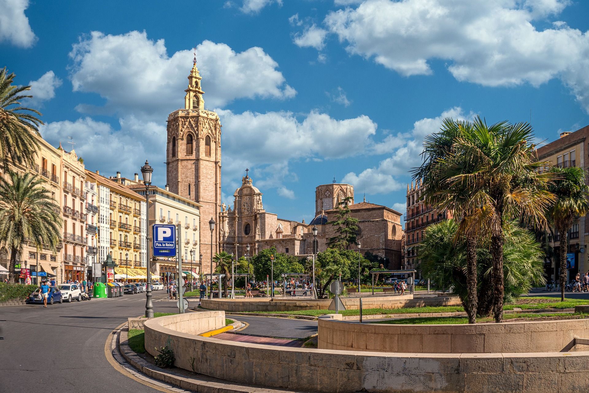 Un campanile storico in pietra si erge sopra una piazza cittadina con palme, edifici circostanti e una strada in una giornata di sole.