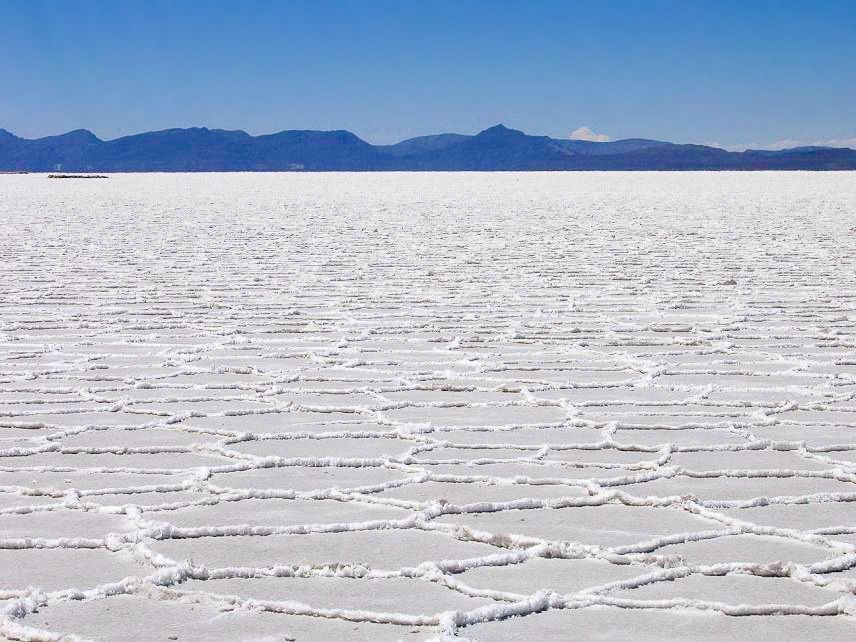 Una vasta distesa salina con naturali motivi esagonali che si estende verso montagne lontane sotto un cielo azzurro e limpido.