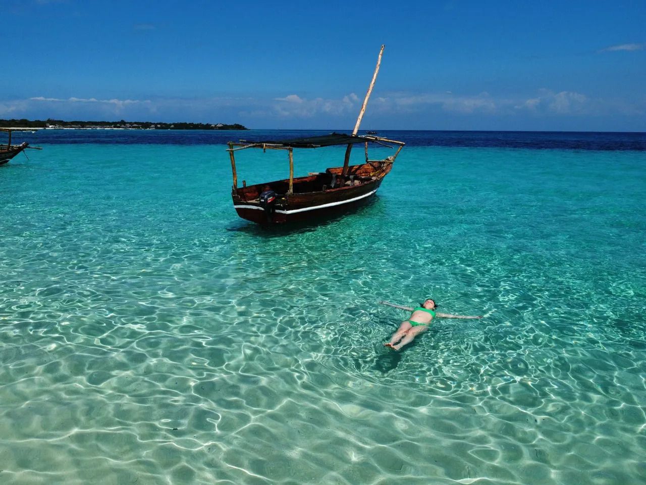 Una donna in bikini verde galleggia sulla schiena in acqua cristallina turchese accanto a una barca tradizionale di legno sotto un cielo soleggiato.