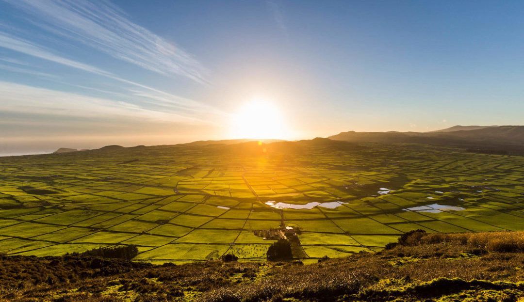 Il sole tramonta su un vasto paesaggio di campi verdi, tingendo le colline lontane di un caldo bagliore dorato sotto un cielo azzurro.