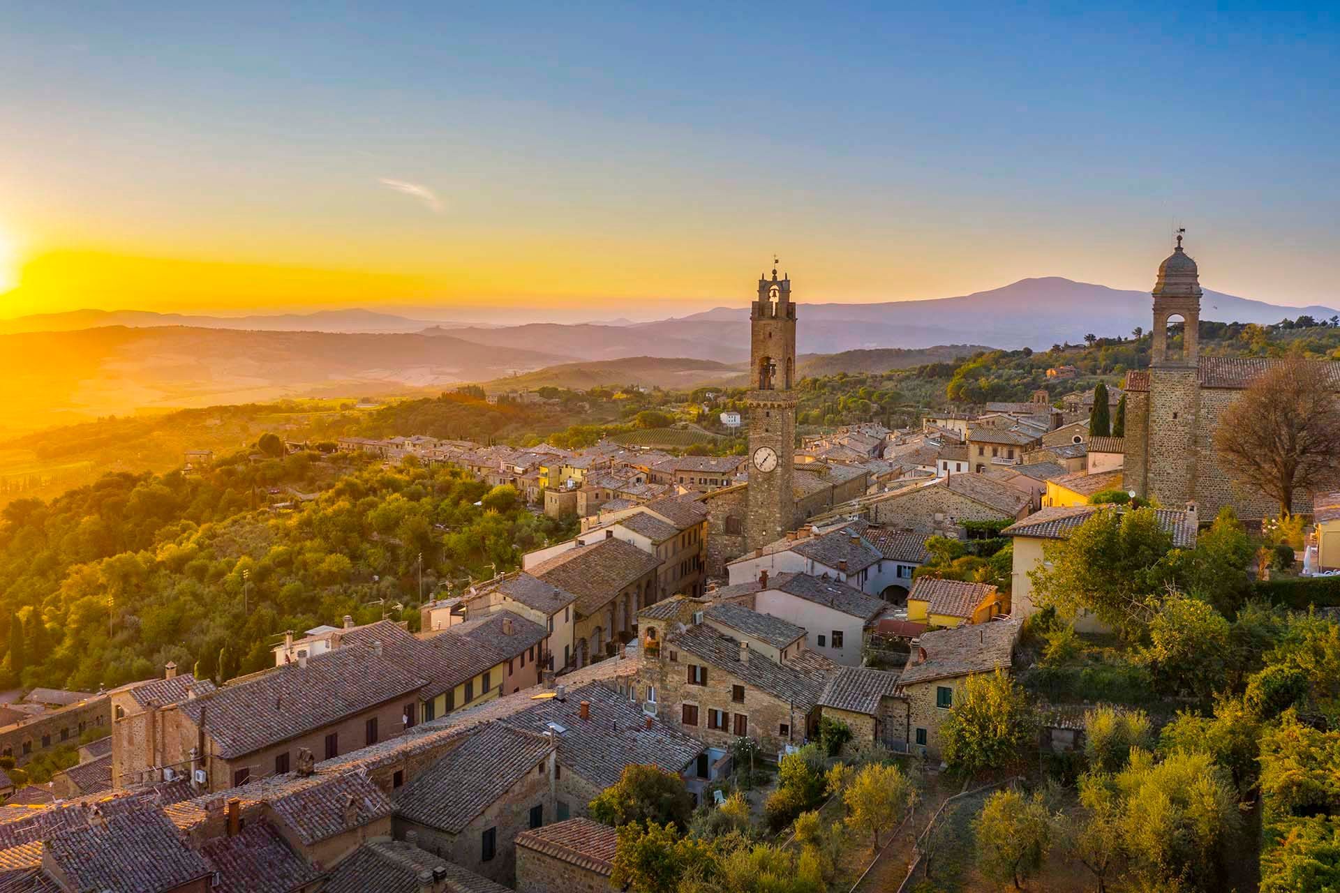 Una vista aerea di una città storica con una torre dell'orologio, circondata da verdi colline e montagne al tramonto.