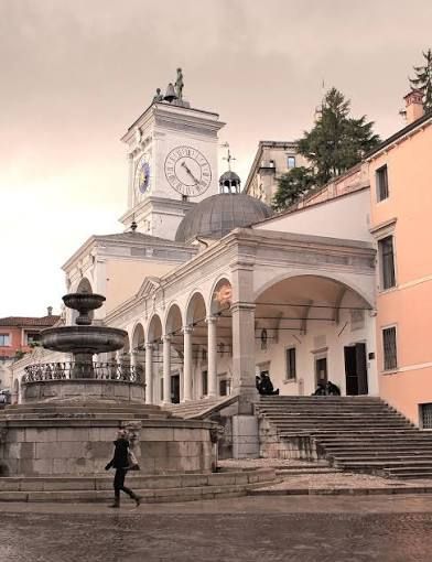 Una persona passa davanti a una grande fontana in pietra in una piazza davanti a un edificio classico con una torre dell'orologio, una cupola e un portico ad archi.