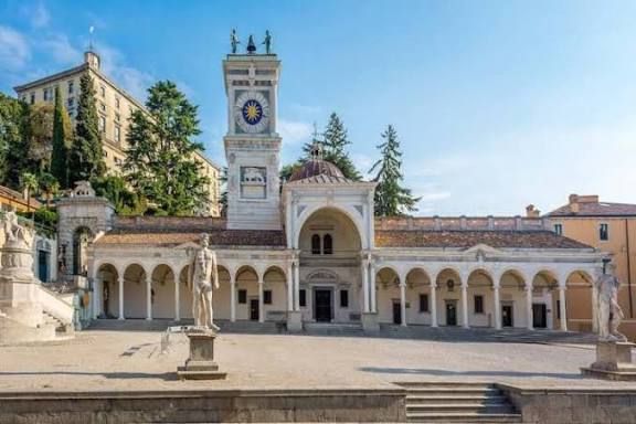 Un edificio storico bianco con una loggia ad archi e una torre dell'orologio in una piazza soleggiata con statue.