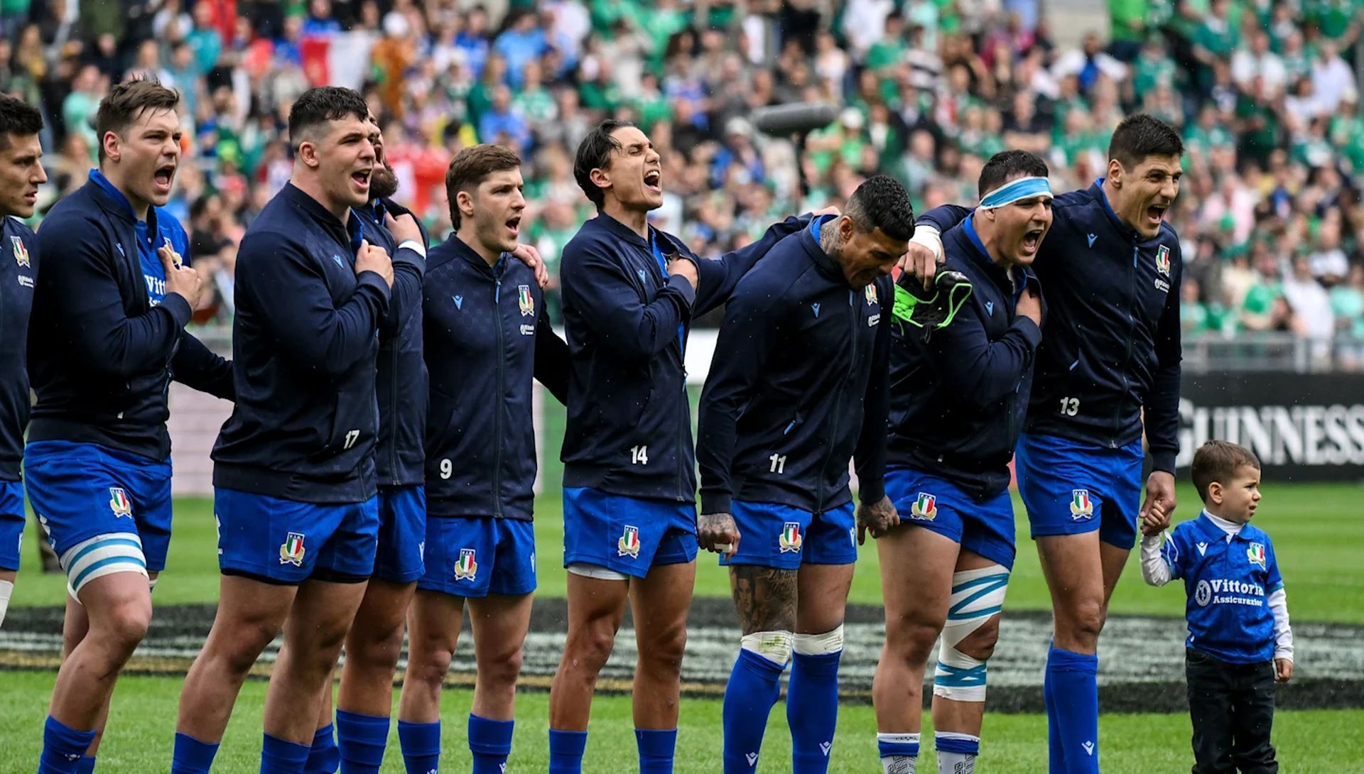 La nazionale italiana di rugby e un giovane ragazzo sono in fila su un campo, cantando con passione prima di una partita.