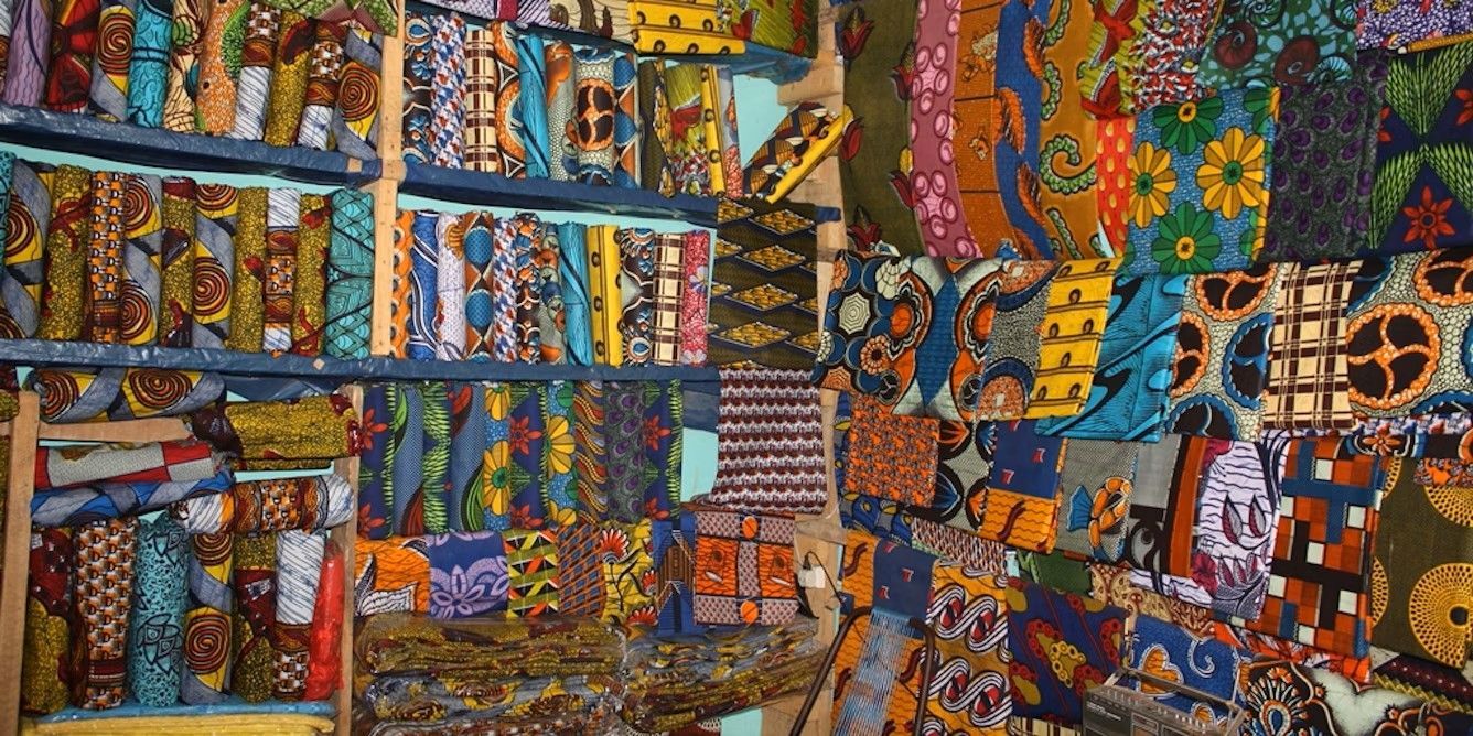Una vasta varietà di tessuti africani wax colorati e fantasia, impilati ed esposti su scaffali di legno in una bancarella del mercato.