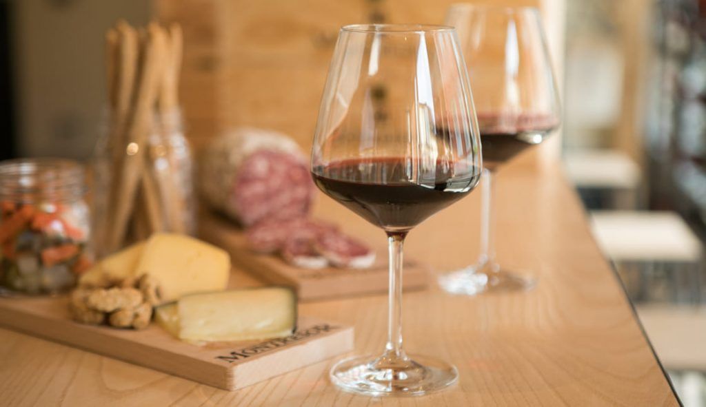 Un bicchiere di vino rosso poggia su un bancone di legno, con taglieri di salumi e formaggi sfocati sullo sfondo.