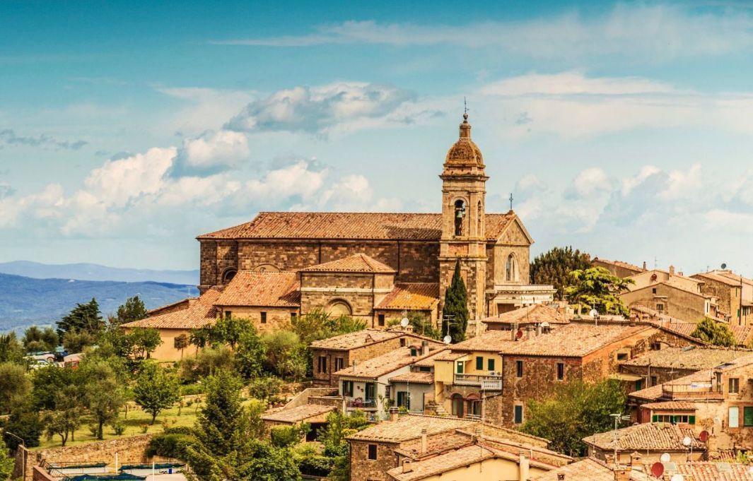 Una chiesa in pietra con un campanile domina un borgo collinare con tetti in terracotta, con le montagne sullo sfondo.