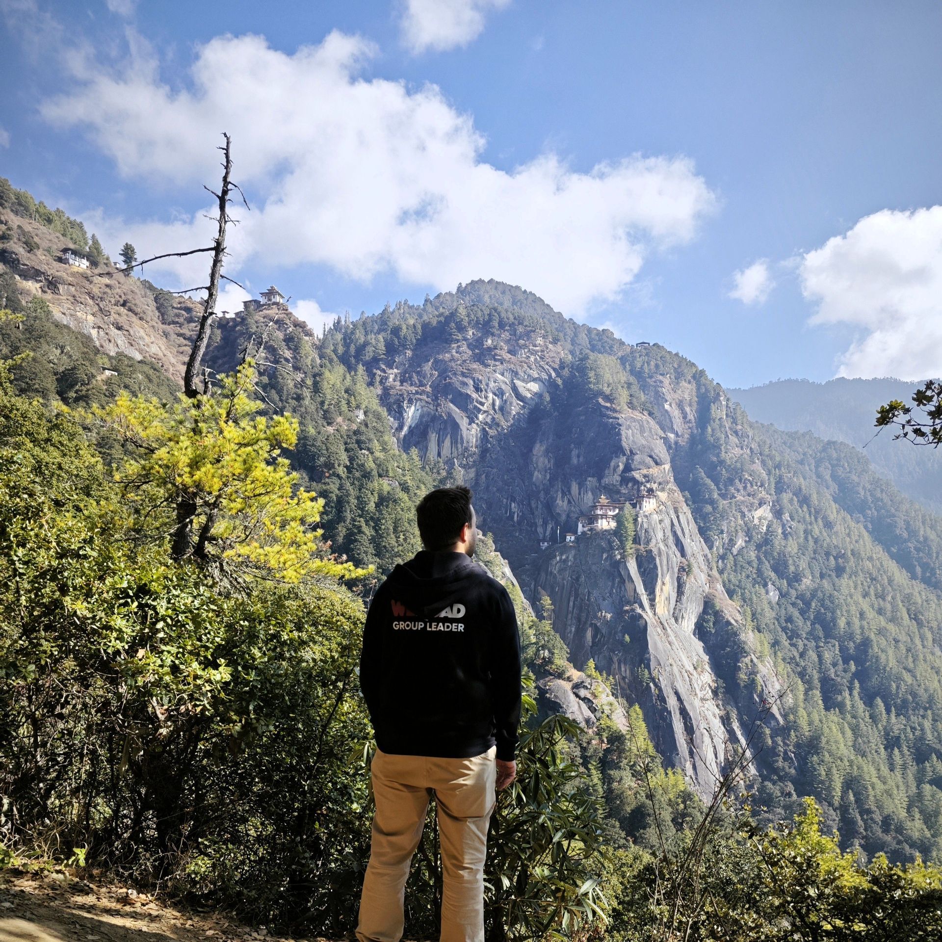 Un Group Leader WeRoad visto di spalle, che ammira un monastero incastonato in una scogliera su una montagna boscosa.