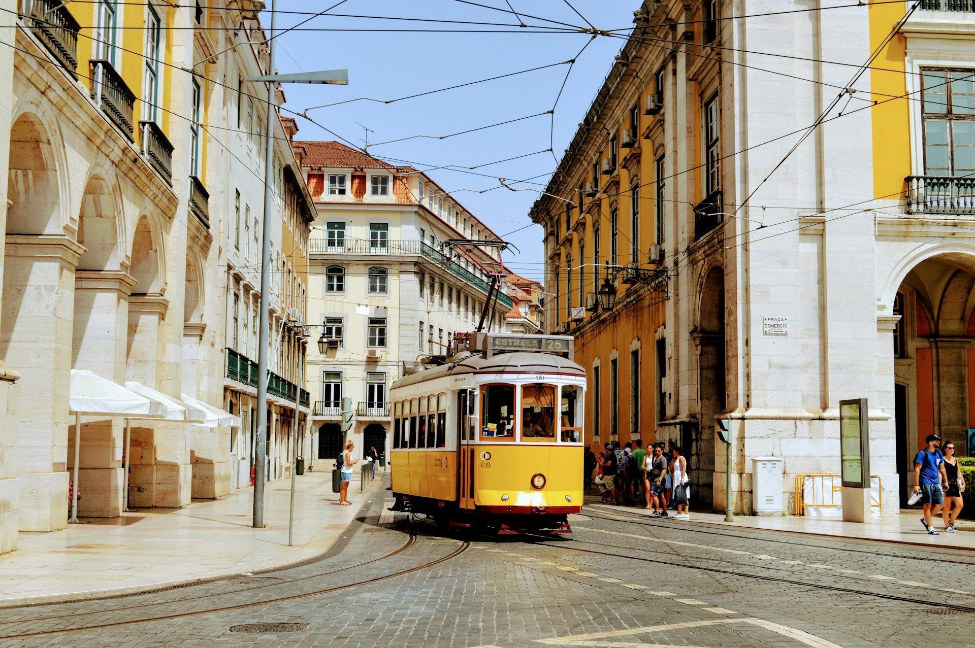 Un classico tram giallo viaggia su una strada acciottolata tra edifici storici con archi, sotto una ragnatela di cavi elettrici.