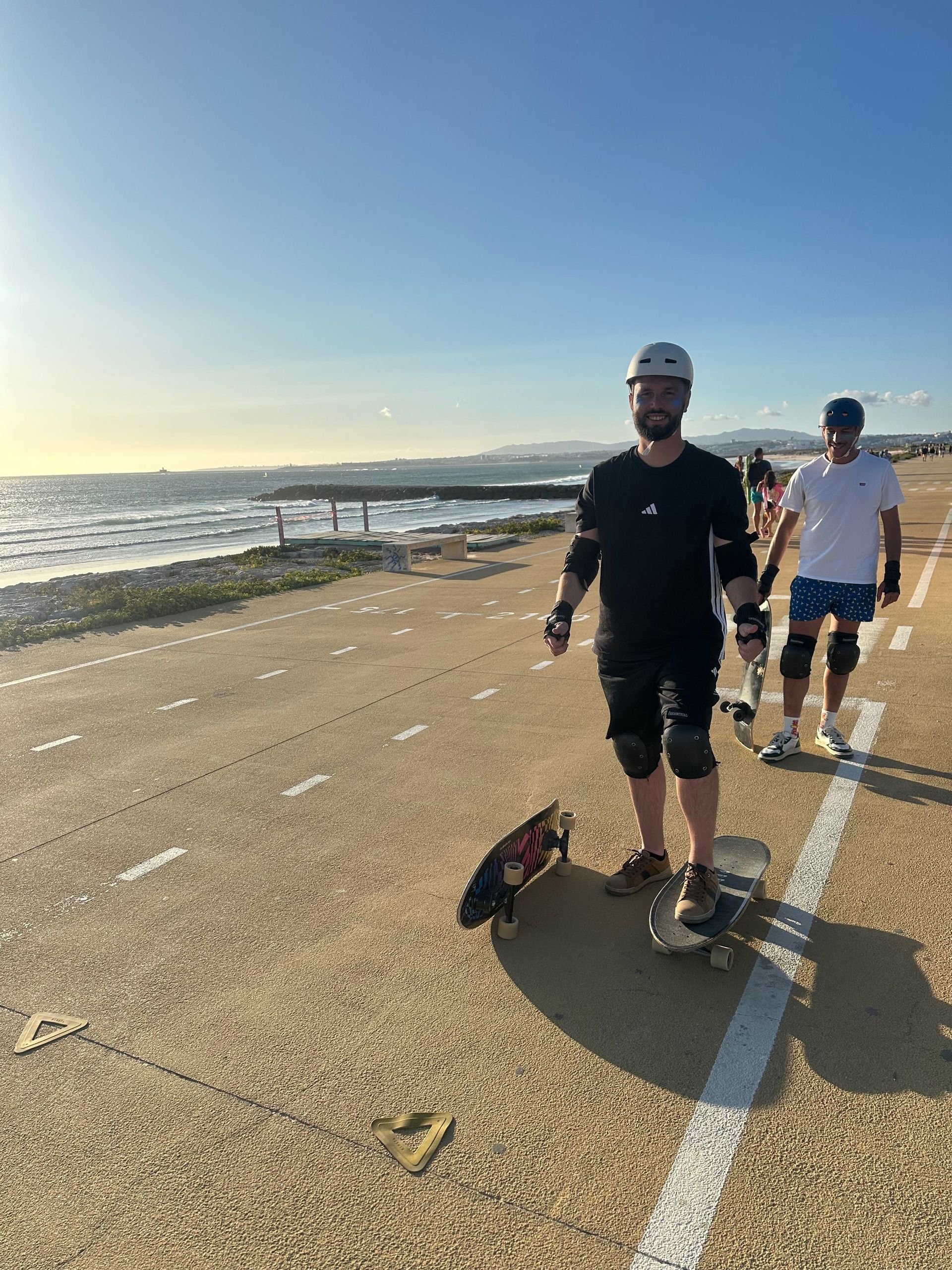 Due uomini di un viaggio di gruppo WeRoad, con caschi e protezioni, sono in piedi con i loro skateboard su un percorso costiero pavimentato al tramonto.