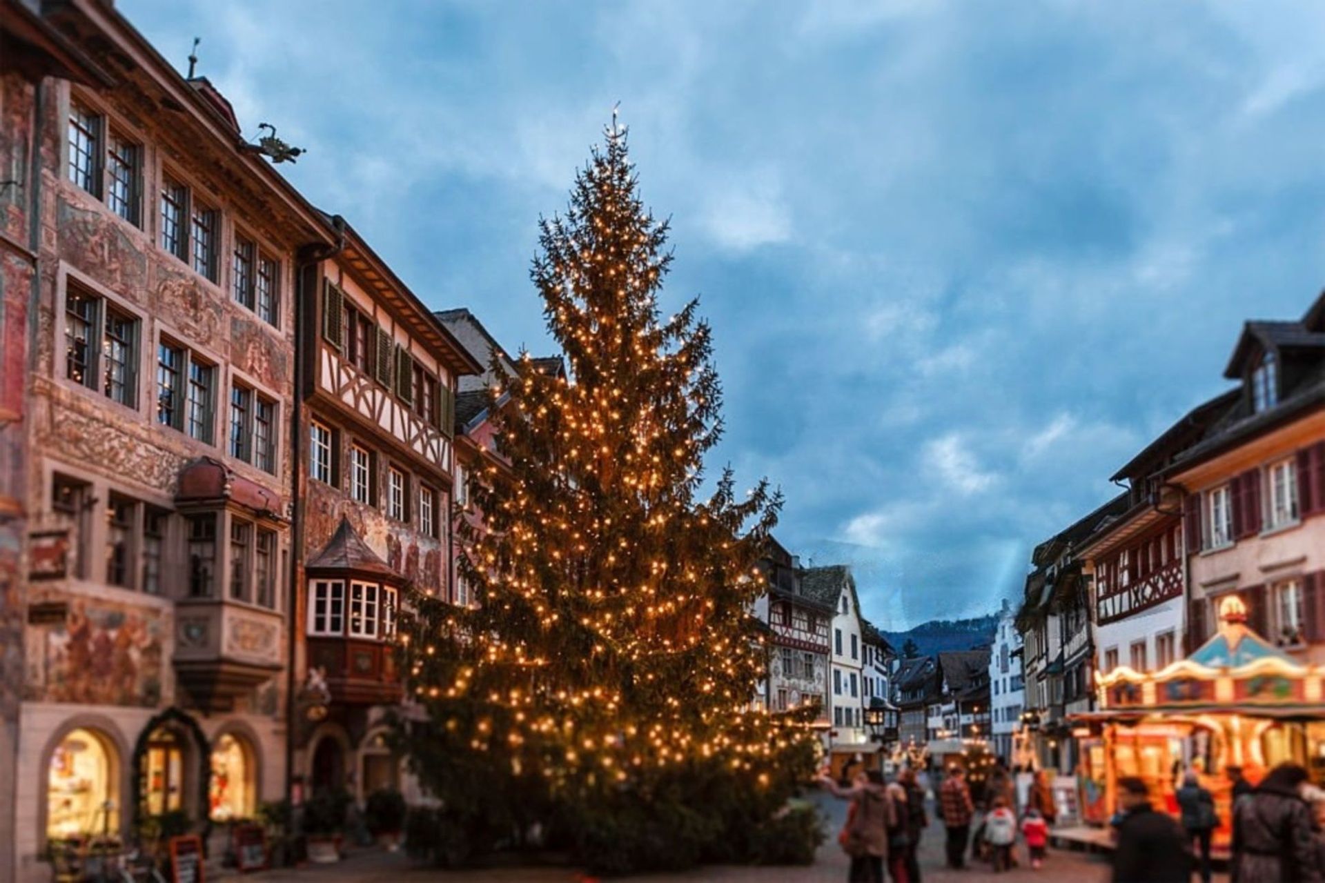 Un grande albero di Natale decorato con luci si erge in una piazza cittadina festosa, circondato da edifici storici e persone al tramonto.