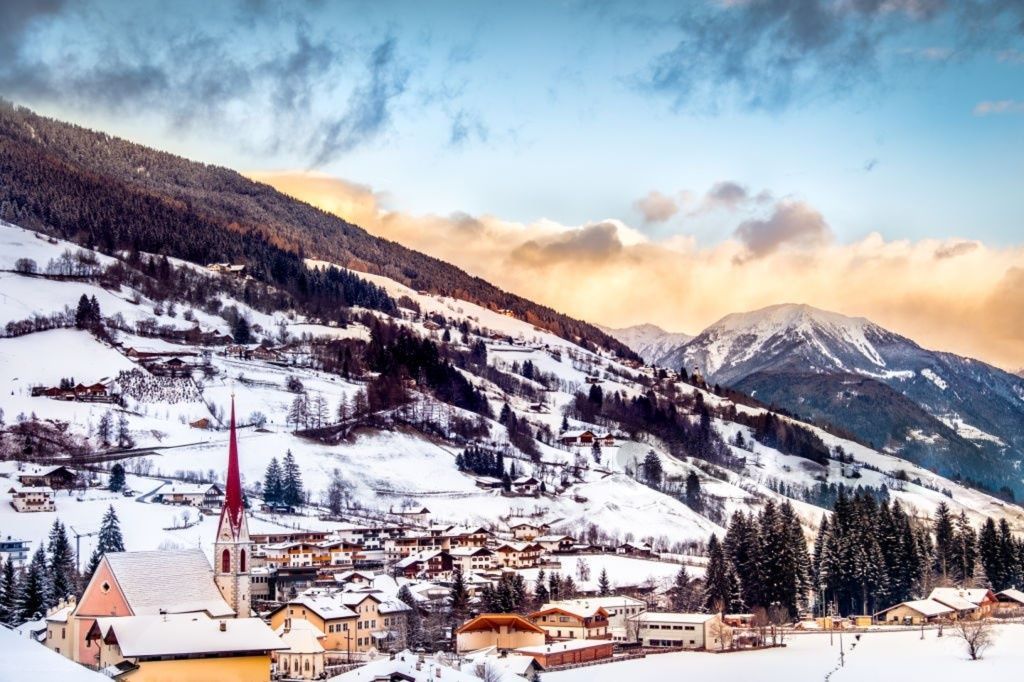 Un villaggio innevato con una chiesa dalla guglia rossa si adagia ai piedi di una catena montuosa boscosa, sotto un cielo nuvoloso al tramonto.
