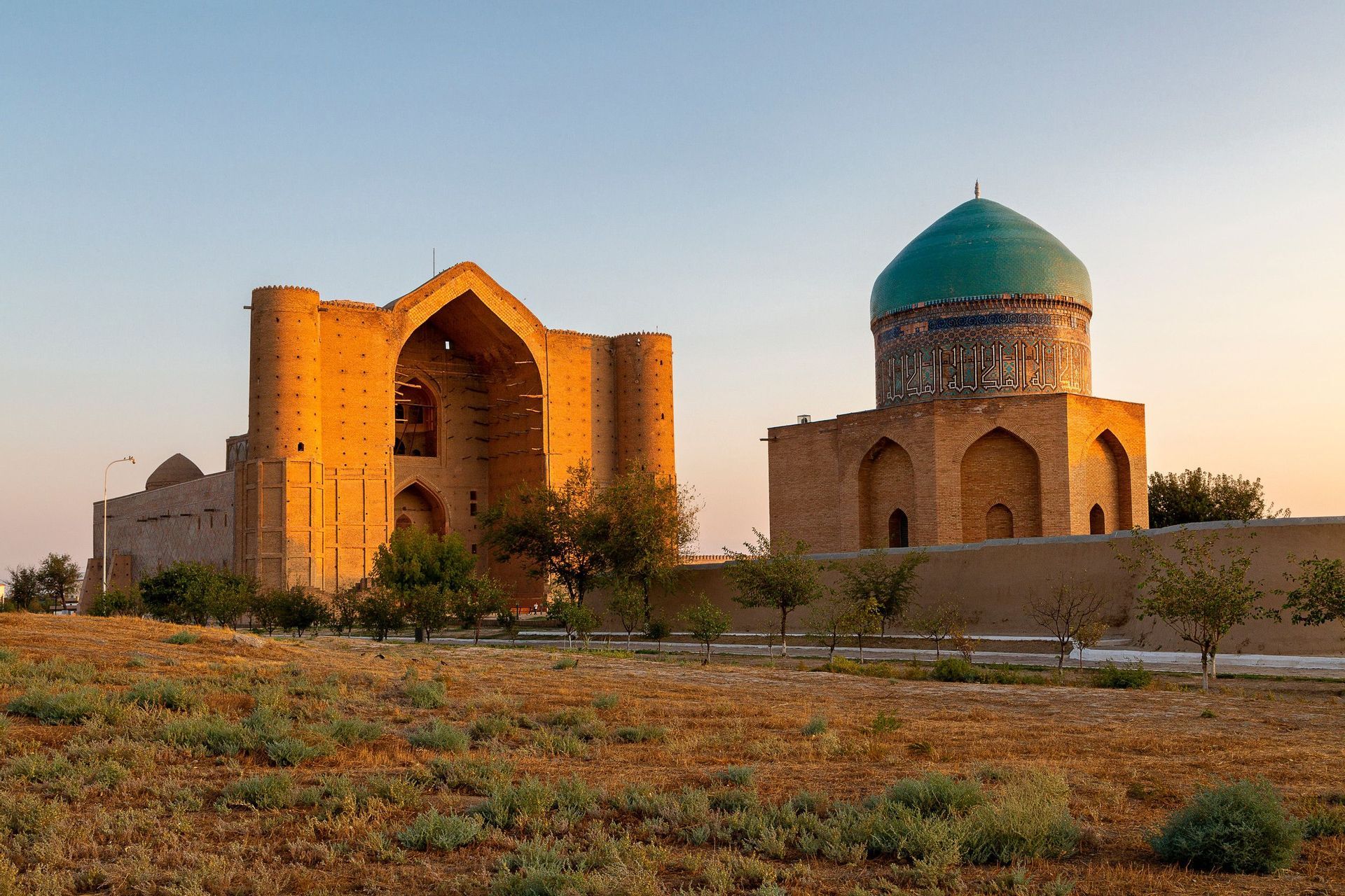 Two historic brick buildings, one with a large turquoise dome, illuminated by golden hour light across a grassy field.